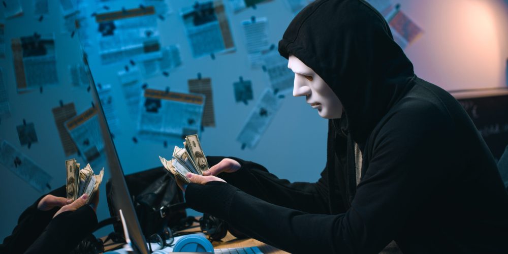 side view of hooded hacker in mask counting stolen money at his workplace